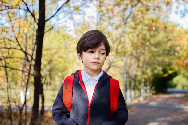 Zamyślony chłopiec z plecakiem szkolnym spacerujący po drodze otoczonej drzewami z obu stron.